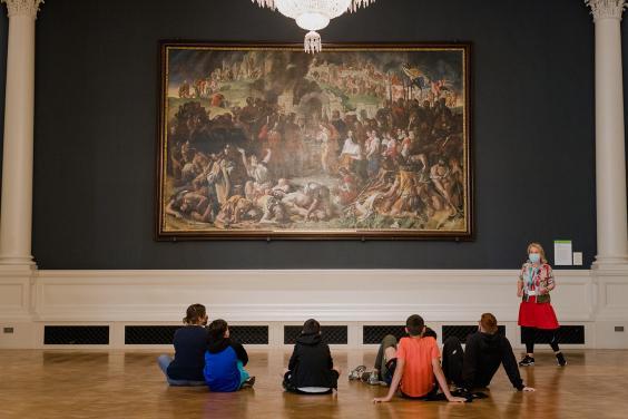 School students sit on floor in front of a large oil painting as a woman wearing a face mask speaks to them