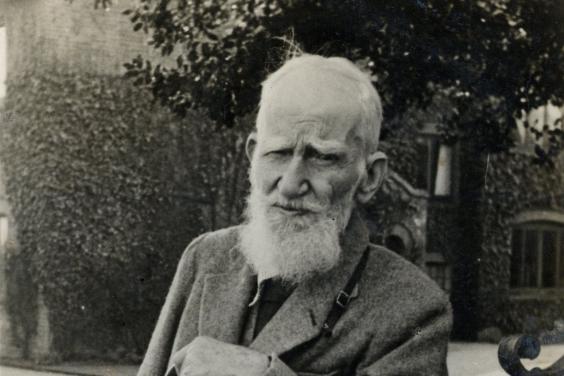 George Bernard Shaw leans on a gate, looking at the viewer. He is wearing a tweed suit, and has a camera around his neck