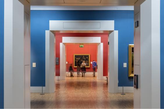 Photograph of a group of people looking at a painting, taken from down a long red and blue corridor.