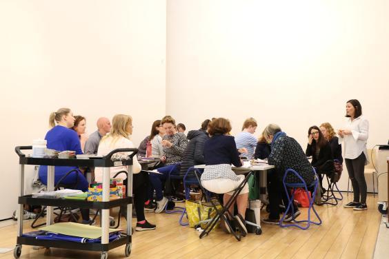 A group of people of various ages and genders sit on chairs around tables in a white room, looking in the direction of a standing woman.