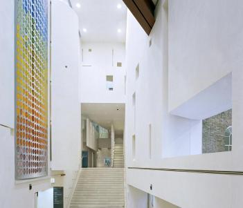 The large interior of a modern white building, with a large hanging tapestry on the left wall made up of a rainbow of spots.