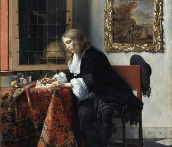 Detail of a man writing at a desk covered in a heavily patterned cloth.