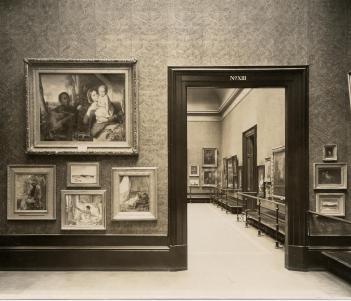 Black and white photo of paintings hung Salon-style on the wall with view through doorway into next gallery space