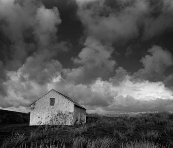 A black and white photograph with a low horizon and expansive cloudy sky, with simple whitewashed cottage standing in a field.