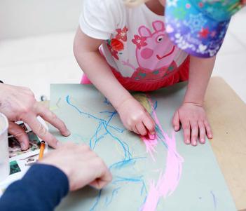 Photo of a young child drawing with an adult's hands stabilising the piece of paper.