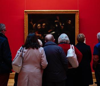 Photo of a group of people, with their backs to the camera, looking at Caravaggio's painting of the Taking of Christ, which is hanging on a red wall.
