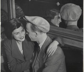 A black and white photograph of a woman and a man sitting side by side at a table, with two half full glasses of wine in front of them. They are turned towards each other, and the woman has her arm around the man's shoulder, and is smiling. Behind them is a large mirror in which we can see them reflected. 
