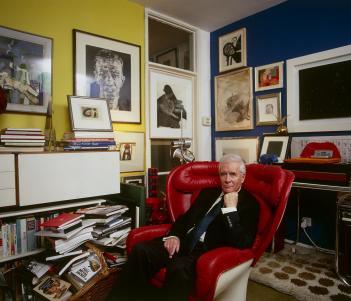 Photo of Alexander Walker in his apartment sitting in a red leather armchair with lots of art hanging on the walls
