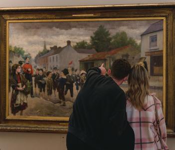 A photograph showing a man and woman standing looking at a large oil painting. Their heads are close together and the man is pointing at something in the painting.