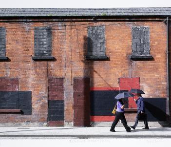 A photograph of a row of red brick houses with the windows and doors boarded up. Two people walk by in opposite directions, both holding umbrellas.