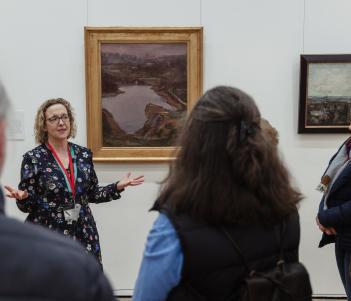 A guide stands in front of a painting, talking to a group of people who are looking at her. 