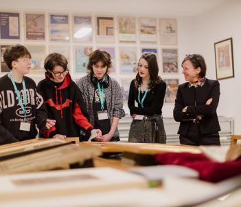 A group of young people standing around discussing prints and painting on a table.