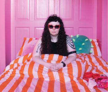 A young girl with long black hair, sits up in a bed with a pink and orange striped duvet, in a pink room.
