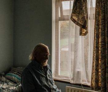 Photograph of an older man with a white beard, sitting in a small room in blue pyjamas