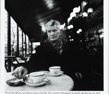 Black and white image of an older male figure sitting at a Parisian cafe, with two coffee cups on the table in front of him.