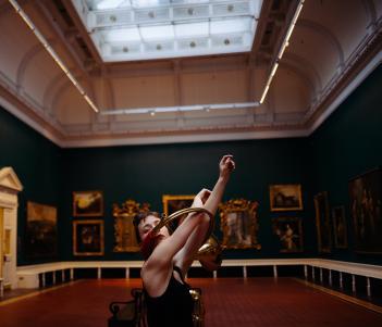 A figure stands in a gallery filled with paintings, arms outstretched through two metal rings.
