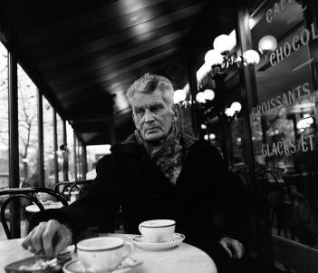 Black and white photograph of Samuel Beckett in later years, sitting at a marble table outside a cafe in Paris with two teacups on the table.
