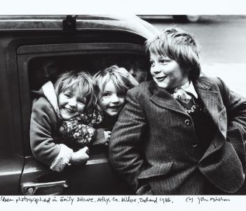 Black and white photograph of three young children laughing in front of a car.