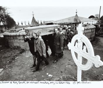 Black and white photograph of pallbearers carrying a coffin in a graveyard, followed by a crowd of mourners.