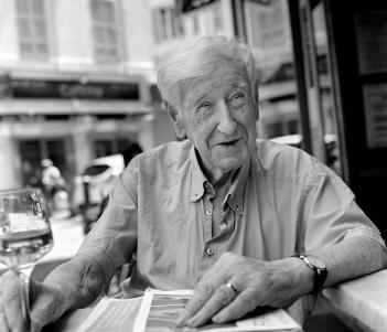 Black and white photograph of an older man with white hair and a short sleeved shirt, sitting at a table in front of a cafe in Nice.