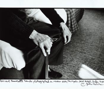 Black and white photograph focusing on an older man's hands as he sits on the edge of a bed, resting his hands on his knees
