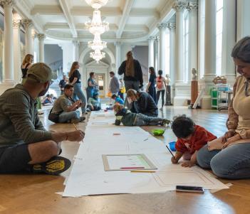 A group of adults and children sit on the floor of a large hall, drawing on a long sheet of paper running the length of the room.