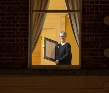 A figure stands at a window, holding a framed certificate.