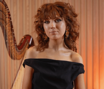 A woman with red hair and wearing an off-the-shoulder black dress stands beside a harp