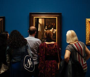 A photograph of people, with their backs to the camera, looking at three gilt-framed paintings on a blue wall.