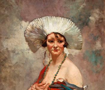 A woman sits wearing a starched white headdress and long beaded necklace. A shawl has fallen off her bare shoulders. 