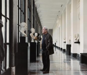 Writer John Banville stands in a corridor of the Prado Museum looking at a row of busts.
