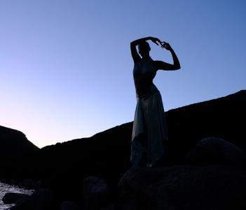 A figure in silhouette stands in a balletic pose, a blue sky and shadowed hills behind them.
