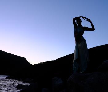 A dancer stands on the banks of a river with her arms linked above her head, silhouetted against a blue sunrise sky. 