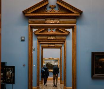 A photograph of a suite of galleries, looking through a series of ornate wooden doorframes. The walls are painted blue, and we can see some paintings hanging. In the distance, some people are walking through the rooms.