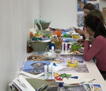 Two people sit at a long table covered in art supplies and materials