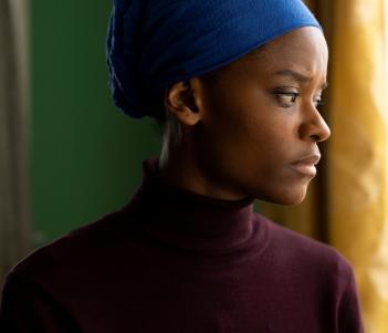 A film still of a young black female figure wearing a burgundy polo neck and a blue headscarf 