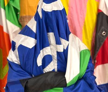 A figure is sitting with their arms folded covered in a large blue and green flag with white writing