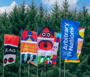 A group of colourful flags, flying in a strong wind, against a backdrop of spruce trees.