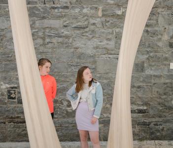 Two young girls standing in front of a stone wall framed by a curved wooden sculpture