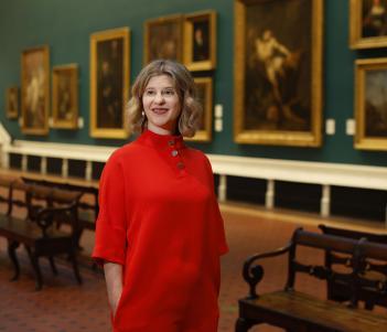 A woman in a red dress stands in a Gallery hung with large oil paintings. 