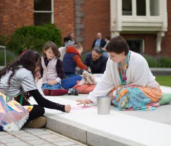 A group of people sit outside on the ground, drawing in crayons and pastels