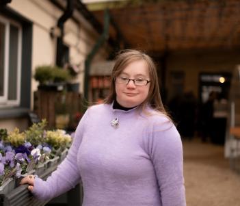A brown-haired woman in a lilac wool jumper stands beside a raised flower bed outside a house with one hand on it