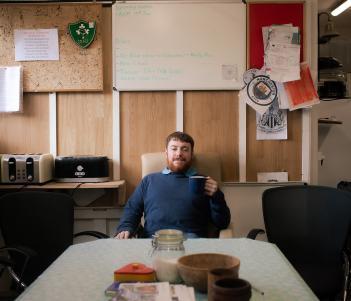 A man in a blue jumper sits at the end of a formica table  under a large yellow sign that says 'Mens Shed'