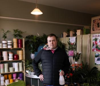 A man in a black puffer jacket and tracksuit stands with one hand on his hip in a room filled with pot plants. In his other hand, a spray gun filled with water. 