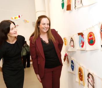 A female figure dressed in black stand beside a female figure wearing a brown suit, looking at artwork created by children.