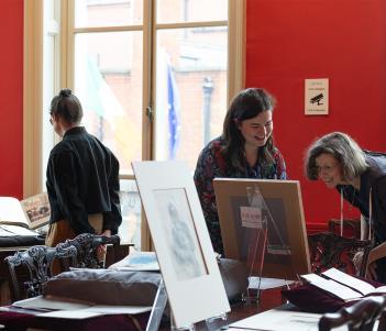 Three female figures in a red room, looking at a collection of drawings, books and other items that have been spread on a table. 