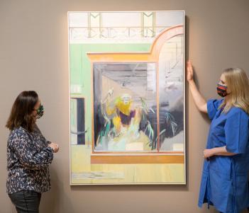 Two women wearing face masks standing either side of a painting that's hanging on a grey wall