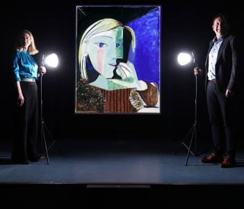 Two figures stand in semi darkness on either side of a large painting by Picasso. In front of both of them is a spotlight.
