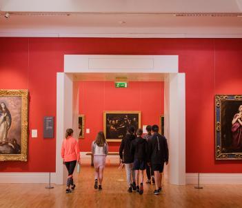 A group of young people walking away through an open doorway in an art gallery with red walls and gilt-framed oil paintings