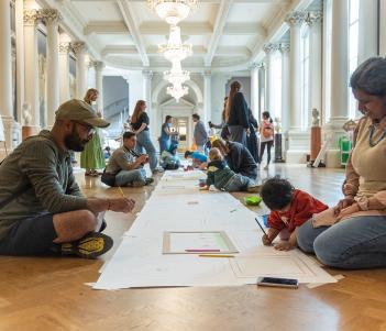 A family draw pictures on a long sheet of paper, which is laid on the floor of a grand room.